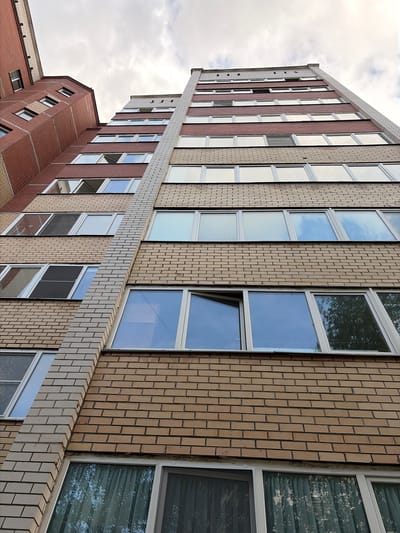 The image shows a multi-story apartment building with a brick facade, observed from a low angle looking upwards. The building features numerous windows, some of which are open. The sky above is partly cloudy. The building's architecture appears to be typical of residential complexes in many Russian cities. Based on the architectural style and common building materials, this scene is likely located in the city of Syktyvkar, Russia. The photograph captures the exterior of the building, highlighting its vertical structure and the repetitive pattern of windows and brickwork. There are no people or vehicles visible in the image. The environment is urban, characterized by the presence of the apartment building under a cloudy sky.