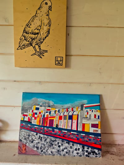 Two pieces of artwork are displayed on a white-painted, horizontally-paneled wall.
The top piece is a light brown, rectangular wooden plaque featuring a line drawing of a bird, possibly a pigeon or dove, standing. The drawing is in black ink. A small logo or signature is present in the lower right corner, consisting of stylized letters.
Below the wooden plaque is a rectangular canvas painting. The painting depicts an abstract cityscape under a blue and partly cloudy sky. The buildings are rendered with bold, bright colors like red, yellow, blue, and white, in a somewhat cubist style. A red and grey pathway is in the foreground, apparently with pedestrians. A small cat is seen in the painting. The location in the city of Свистуха (кузяевский С/о), Russia is not evident from the image.