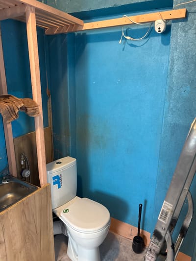 This image depicts the interior of a small, unfinished room or enclosure, likely a bathroom or a utility space. The walls and floor are painted a vibrant blue. In the foreground, a wooden structure, possibly a bench or shelving unit, is partially constructed. A beige towel hangs from the wooden frame.
To the right of the frame, a white toilet is installed. It appears to be a compact model, possibly designed for a smaller space. The toilet has a brand logo with blue lettering that reads "THEET" and a green circular sticker with a leaf design on the tank. Adjacent to the toilet, a white tank, possibly a water heater, is visible.
Further to the left, a small white security camera is mounted on a wooden post. It is connected by white wires. In the upper right quadrant of the image, a silver ladder is leaning against the blue wall. A barcode label is attached to one of the ladder's rungs.
The scene appears to be in an indoor setting, with artificial lighting. There are no people visible in the image. The overall impression is of a space undergoing renovation or construction. The specific location of Vileyka, Belarus, is noted.