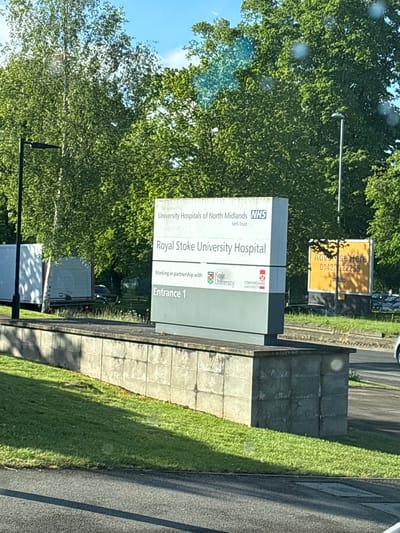 The image shows a sign for Royal Stoke University Hospital, located in Newcastle-under-Lyme, United Kingdom. The sign includes the NHS logo and the logos for Keele University and Staffordshire University, indicating partnerships. The sign also directs people to "Entrance 1." Another sign for advertising is also seen in the background. The sign is surrounded by green trees and grass. A road is visible to the right of the sign. A pole holding up the signs is on the left side. Other vehicles are parked in the background near the main sign.