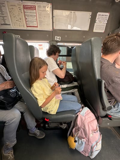 In the city of Verkh-Sosnova, Russia, this image captures the interior of a passenger bus. Two individuals are seated in gray bus seats. In the foreground, a young girl with blonde hair, wearing a yellow t-shirt and blue jeans, sits facing right and appears to be interacting with a pink object in her hands. To her right, a pink backpack with a yellow fuzzy charm is placed on the floor. Behind her, a young man with curly brown hair, wearing a white t-shirt, is seated facing forward and looking down. Further behind them, another passenger is partially visible. The bus interior features informational posters and signage on the walls. The overall environment suggests a public transportation setting.