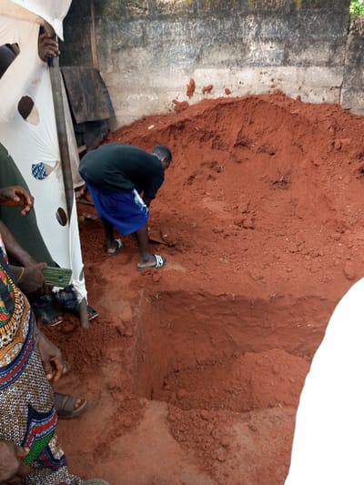 The scene is set in Oko Town, Nigeria. A male subject is observed actively digging a rectangular pit in reddish-brown soil. He wears a black t-shirt, blue shorts, and white-and-black striped slides, bending forward over the excavation. A substantial mound of excavated reddish-brown earth is piled directly behind him, against a weathered grey concrete block wall. To the left, several individuals are partially visible, obscured by a torn white fabric tarp supported by poles. One individual's left hand is visible holding a dark green smartphone, while another's lower body shows a colorful patterned fabric garment. The ground surface consists entirely of reddish-brown earth.