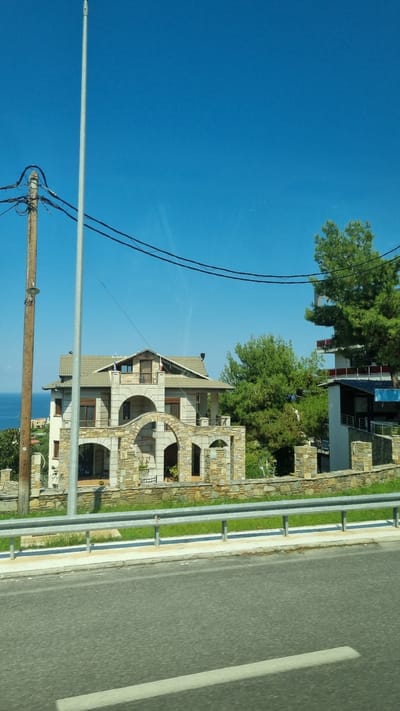 The image depicts a multi-story stone house with arched entrances and balconies, situated on a hillside overlooking the sea. The house features a light-colored tiled roof and stone accents on its facade. It is surrounded by green vegetation and mature trees. In the foreground, a metal guardrail separates the property from a paved road. Power lines are visible strung across the bright blue sky, supported by a wooden pole on the left side of the frame. The scene appears to be located in Neos Panteleimonas, Greece, as indicated by the context. The overall impression is of a scenic, residential area with a view of the coastline.