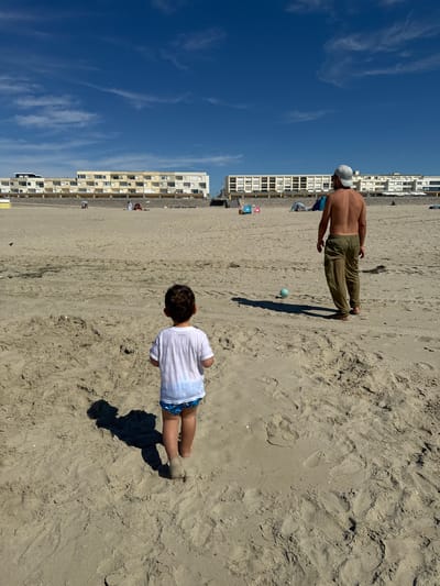 The image depicts a beach in Berck, France. In the foreground, a young child wearing a white t-shirt and blue patterned shorts stands facing away from the camera, their shadow cast on the sand. Further back, an adult male, shirtless and wearing khaki pants and a white baseball cap, also faces away from the camera, looking towards a blue and green striped ball on the sand. In the background, a long, multi-story building complex runs along the coastline, with a strip of sand and vegetation separating it from the beach. The sky is a clear blue with scattered white clouds. Several other people and beach umbrellas are visible in the distance near the buildings. The overall scene is a sunny day at the beach.
