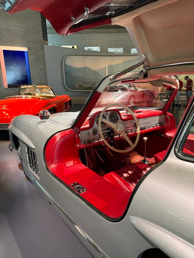 The image shows the interior of a classic Mercedes-Benz 300 SL Gullwing coupe at the Mercedes-Benz Museum in Stuttgart, Germany. The car's iconic gullwing doors are open, revealing a striking red interior with leather seats, dashboard, and door panels. A white steering wheel with a beige rim is prominent in the foreground. In the background, another red Mercedes-Benz convertible is visible, along with a display of artwork on a wall. Two individuals, a man and a woman, are seen in the distance, looking at the exhibits. The museum environment features clean, modern architecture with concrete walls and a large panoramic window displaying a landscape scene. The overall impression is one of a meticulously preserved automotive exhibit in a well-lit museum setting.