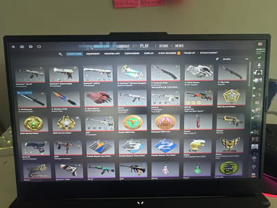 The image shows a computer screen displaying the inventory section of the video game Counter-Strike 2 (CS2). The "Inventory" tab is selected at the top of the screen, and various in-game items are displayed, including weapons (AK-47, Desert Eagle, USP-S), knives (Butterfly Knife, Flip Knife), gloves, service medals, coins, charms, and stickers. The items have different skins and rarities. The game interface elements, such as the search bar, filter options (Quality), and item categories (Everything, Equipment, Graphic Art, Containers, Display, X-Ray Scanner, Trade Up, Steam Market), are also visible. The computer is a laptop, and there are sticky notes on the wall behind it.

This image does not contain information about any location in the city of Rüsselsheim am Main, Germany.