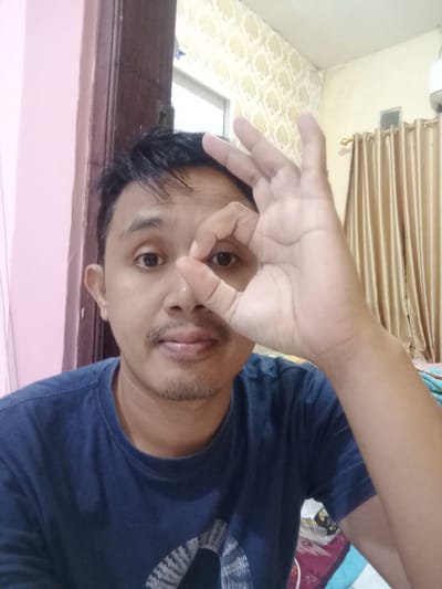 In an indoor setting in Brangsong, Indonesia, a single adult male is depicted from the chest up, facing the viewer directly. He has short black hair, dark eyes, a light mustache, and stubble on his chin and jawline. He is wearing a dark blue short-sleeved t-shirt with a light-colored graphic design on the lower front. His left hand is raised to the level of his face, forming an "okay" gesture with his thumb and index finger, positioned over his left eye. The background consists of a pink wall and a dark brown door frame on the left. To the right, a wall features light-colored wallpaper with a repetitive gold or yellow pattern. Visible elements include a window opening above the subject's head, gold-colored curtains drawn on the right, a white air conditioning unit mounted high on the wall, and a multicolored patterned fabric in the lower right quadrant of the image.