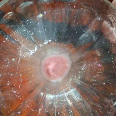 This is a close-up, overhead shot of the inside of a clear glass bottle, possibly a soda bottle, with a pinkish, creamy substance at the bottom, possibly a beverage or food item. The glass has visible ridges that radiate from the center. The liquid or substance is swirling and appears to have some texture, possibly containing small bits. There are numerous small air bubbles and condensation clinging to the inside of the glass, suggesting it might be cold. Scattered throughout the glass are small, colorful flecks, including pink hearts and some yellow and green specks, which could be glitter or decorative elements. The lighting is somewhat diffused, creating reflections on the glass surface. The background visible through the glass is a warm, dark orange and black, possibly indicating the color of the beverage or an object placed behind the bottle. No people are directly visible, but a hand or finger is faintly visible near the top left edge of the frame, possibly holding the bottle. There is no visible text. The overall impression is of a close-up, abstract view of a drink or confection within a bottle, with added decorative elements.
