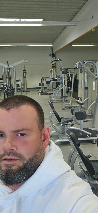 The image depicts the interior of a gym located in Rietberg, Germany. In the immediate foreground, occupying the lower left portion of the frame, is a male subject. He has a short buzz cut, a dark beard, blue eyes, and visible reddish coloration on his cheeks. He is wearing a white hooded sweatshirt.

The background shows an array of professional gym equipment, including various weight training machines with metal frames and black padded seats and backrests. Examples include cable machines, selectorized weight stacks, and a recumbent-style cardio machine. The floor is light-colored, and the walls are a pale hue. The ceiling is a corrugated industrial style with multiple linear fluorescent light fixtures providing illumination. A vertical banner displaying the partially visible word "NEWFIT" is present in the mid-background, suggesting the name of the fitness facility. The male subject is looking directly towards the camera.