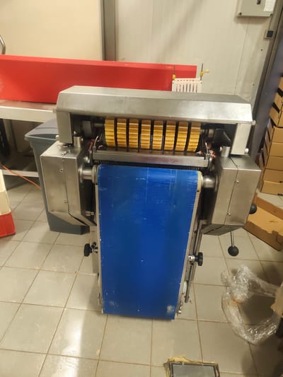 The image shows a commercial food processing machine, likely a slicer, in an industrial kitchen or food production facility. The machine is made of stainless steel and has a prominent blue conveyor belt. Above the belt, a set of rotating blades or rollers are visible, designed to cut food items. The machine is situated on a tiled floor, and in the background, a large red food-grade container is visible on a stainless steel counter. Cardboard boxes are stacked to the right, suggesting storage or incoming materials. The overall setting appears to be a clean, functional workspace, typical of a commercial food environment. There are no people visible in the image. The lighting suggests an indoor setting with artificial light. No text is clearly legible on the machine or its surroundings.