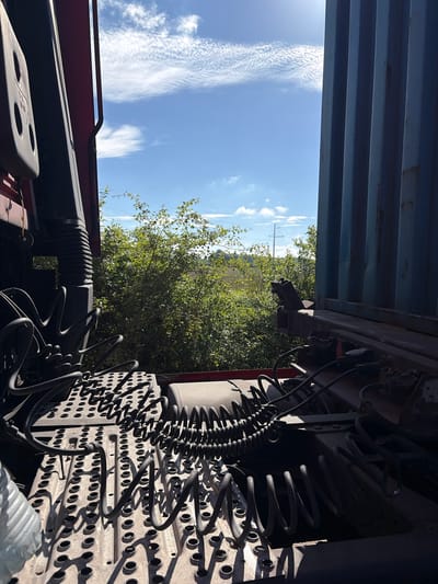 The image shows a view from the inside of a truck cabin looking outwards. In the foreground, a metallic platform with numerous circular indentations is visible, along with coiled black cables. To the left, a portion of the truck's interior, including a dark, ribbed pipe and red paneling, is present. To the right, the blue corrugated wall of a shipping container is partially visible, suggesting the truck is a semi-trailer. The background features lush green trees and bushes under a bright blue sky with wispy white clouds. Power lines and a utility pole can be seen in the distance. The scene appears to be outdoors in a rural or semi-rural area. No people or specific actions are depicted. The location is identified as Zarech'e, Russia.