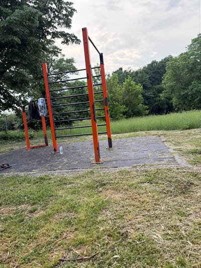 This image captures an unoccupied outdoor calisthenics park in Dolno Sahrane, Bulgaria, observed on an overcast day. The scene is dominated by a weathered orange and black metal exercise structure featuring multiple pull-up bars and a vertical ladder section. Rust is evident on the orange support posts. A black garment and a white-patterned fabric item are draped over separate horizontal bars. A clear plastic water bottle with a white cap stands upright on the dark grey square tiles forming the exercise surface, which is covered with dry grass clippings. To the left foreground, two black and red push-up handles rest on the tiles. Surrounding the tiled area is green grass, appearing recently mown immediately adjacent to the tiles, and transitioning to longer, untrimmed grass further back and to the right. A small stick lies on the grass in the foreground. The background consists of a dense treeline with varied green foliage, and the sky above shows scattered white and grey clouds. No people are visible.
