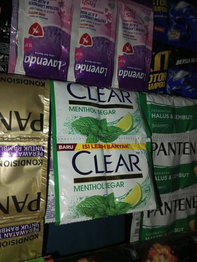 This is a close-up, eye-level, indoor shot of a collection of personal care product sachets and packages, likely displayed for sale in a small shop or market stall in Turumame, Indonesia. The lighting is dim, suggesting the environment might be a dimly lit store or an evening setting.

The most prominent items are sachets of Clear shampoo, featuring imagery of mint leaves and lime slices, indicating a "Menthol Segar" (Fresh Menthol) variant. Two such sachets are stacked vertically in the center of the frame. To the left and right of these, there are sachets of Pantene hair conditioner and shampoo, also in various packaging colors, one gold and one white with green accents.

Further to the left, there are sachets of what appears to be "Lavenda" lotion, with floral patterns and text in Indonesian. In the background, some larger packages of "X.TOR" shampoo are visible, predominantly in blue and yellow packaging.

The overall impression is one of a retail display of commonly used toiletries, arranged somewhat haphazardly. The sachets are clean and appear to be new. There are no people or actions depicted in the image. The focus is entirely on the products themselves.