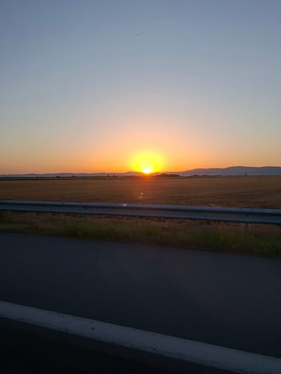 The image captures a sunset over a rural landscape, likely in or near Загорци, Bulgaria, given the context. The scene is dominated by the warm hues of the setting sun, which casts a bright orange glow across the sky and illuminates a vast, harvested field of dry, golden-brown crops. In the distance, a range of low, dark hills forms the horizon, punctuated by what appear to be man-made structures, possibly water towers or silos, silhouetted against the fading light.
In the foreground, a paved road runs diagonally across the lower portion of the frame, with a metal guardrail separating it from the field. The road is dark, indicating it might be asphalt, and there is a white line painted along its edge. The guardrail is silver and shows signs of wear. The sky transitions from a bright orange near the horizon to a soft, pale blue at the top of the image, with a faint, thin contrail visible in the upper right quadrant. The overall impression is one of tranquility and the end of a day in a natural, open environment.