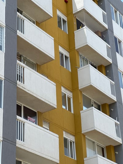 The image shows a close-up, eye-level view of the exterior of a modern apartment building in Barranquilla, Colombia. The building features a striking facade with alternating sections of gray and a vibrant yellow. Numerous balconies, made of white concrete with white railings, project from the building. These balconies are stacked vertically, creating a rhythmic pattern. Some balconies have netting installed for safety.
Windows with white frames and blinds are visible on various floors. One window on a lower floor, on the yellow section of the building, displays a red "SE VENDE" (For Sale) sign. The sign is rectangular and has white text that includes "APARTAMENTO" and a phone number, "300 6213950".
The architecture suggests a contemporary residential building in an urban setting. The lighting indicates it is daytime, with sunlight illuminating the facade. There are no people visible in the image. The overall impression is one of residential architecture with a colorful design.