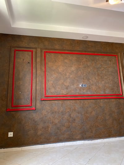 The image shows an interior wall in a building located in Baraki, Algeria. The wall has a textured brown finish and is adorned with decorative moldings that are outlined with red tape. Two framed sections are visible on the wall: a taller, narrower one on the left and a wider, rectangular one to its right. Both frames are also outlined with red tape, suggesting an ongoing renovation or decorative process. Electrical outlets are present on the wall; one is visible within the larger frame, and another is located near the bottom left corner of the wall. The ceiling is white and features recessed lighting. The floor is tiled with light-colored square tiles. The scene appears to be an indoor residential or commercial space undergoing some form of finishing work.