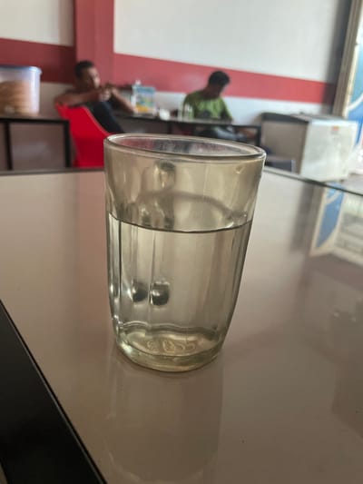 The image shows a close-up of a glass of water on a reflective surface, likely a table. The background is blurred but suggests an indoor setting with people. To the left, a person is sitting in a red chair, facing away from the camera. Further back, another person is seated, wearing a green shirt. The environment appears to be a casual eatery or cafe. The overall lighting is bright. There are no visible signs or indicators to suggest the location is Cotseutui, Indonesia.