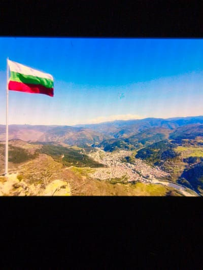 The image shows a panoramic view from a high vantage point, likely a mountain peak, in the city of Devin, Bulgaria. The Bulgarian flag is prominently displayed on a flagpole in the foreground. In the midground, a vast landscape unfolds, revealing a city nestled within a valley, surrounded by rolling hills and mountainous terrain. A winding river can be seen tracing its path through the valley, adding to the scenic beauty of the location. The sky above is clear and blue.