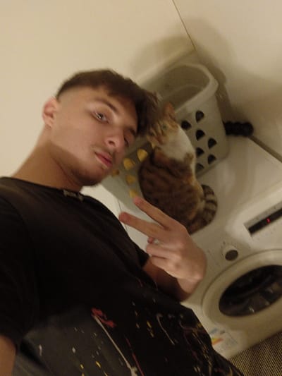 The image depicts an indoor scene in Blaye, France, featuring one male subject and one domestic cat. The male, positioned in the foreground and appearing to take a selfie, is a young adult with short hair faded on the sides and longer on top, light eyes, and light stubble. He wears a dark t-shirt with multicolored paint splatters on the lower front. His right hand (viewer's left) is raised, displaying a "peace" or V-sign. The cat, a tri-colored feline with brown, orange, and white fur, is seated in the midground on top of a white front-loading washing machine. It is looking upwards and slightly to the left, towards the male subject. Scattered next to the cat on the washing machine are several small, yellow, rounded objects. Behind the cat and to its right is a light-colored laundry basket with perforated half-circle patterns. The surrounding walls are plain and light-colored.