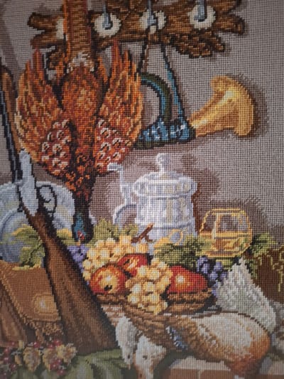 This is a close-up, eye-level, medium shot of a cross-stitch tapestry. The tapestry depicts a still life composition, possibly a hunting or harvest theme, with various objects arranged together.
Dominating the upper left is a large, ornate bird with intricate feather detail in shades of orange, brown, and white. To its right, a golden horn-like object juts out. Below these, a light blue object with black detailing hangs.
In the central area, a white, detailed pitcher or jug with a lid and handle is prominent. To the right of the pitcher, a yellow, rectangular object with dark lines, possibly representing a cart or wagon, is visible.
The foreground is filled with a basket overflowing with fruit, including apples in shades of red and orange, and bunches of grapes in purple and yellow. To the left of the basket, a portion of a rifle or gun is visible, with its stock and barrel. Behind the gun, a brown bag or pouch with a flap closure is present. A white object, possibly a bird or small animal, is positioned in the lower right corner, partially obscured by the fruit basket. Green foliage and other smaller decorative elements are scattered throughout the scene. The background is a neutral, woven texture in shades of gray and beige, characteristic of cross-stitch fabric.
There are no people visible in the image. The objects are static and arranged in a decorative manner. The environment is an indoor still-life as depicted in a textile art piece. There is no information in the image to associate it with Nordhausen, Germany.
