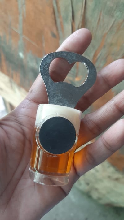 A hand is holding a novelty bottle opener designed to resemble a glass of beer. The bottle opener is made of metal and has a clear plastic base filled with amber liquid to represent beer, topped with a white material simulating foam. A black circular emblem is affixed to the side of the "beer glass." The background is a textured, wooden surface. The image was taken in Kawit, Philippines.