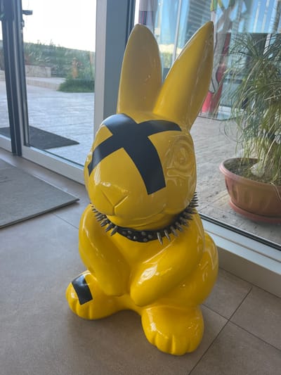 The scene is an interior view from the A. S. Hotel & Residence in Galatina, Italy, featuring a large, glossy yellow rabbit sculpture. The sculpture is positioned on a light-colored tiled floor, facing slightly right, directly adjacent to a glass exterior wall. It exhibits a black 'X' mark across its face where eyes would typically be and wears a black collar adorned with silver-colored spikes around its neck. A black band is visible on its left foot. To the immediate left of the sculpture, a grey rectangular mat lies on the floor next to a glass door frame. To the right, a potted plant with green foliage, contained within a terracotta-colored pot and saucer, is situated beside the glass paneling. Through the glass, an outdoor paved area, landscaped sections with green vegetation, and distant building structures are visible under bright daylight. No individuals are present in the image.