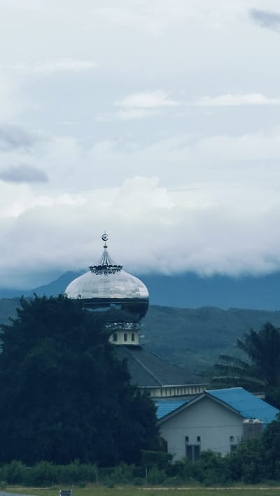 This vertical shot showcases a mosque dome and its surrounding natural landscape in Lambarotunong, Indonesia, under an overcast sky. The focal point is a large, gleaming silver mosque dome, prominently positioned slightly above the center of the frame. It is topped with a distinct crescent moon and star finial. Below the dome, parts of the mosque's structure with a dark roof are partially visible.
Lush, dark green foliage, primarily trees, occupies the midground and foreground, partially obscuring the lower sections of the mosque and other buildings. To the right of the mosque, a small white building with a vibrant blue roof is nestled amidst the greenery. In the background, layers of rolling hills and mountains rise, partially obscured by thick, low-hanging clouds that stretch across the horizon. The sky is dominated by a dense covering of white and grey clouds, indicating a cloudy or possibly stormy day, diffusing the light softly across the scene.
No human figures or explicit activities are visible, lending a quiet and tranquil atmosphere to the image. The scene captures a harmonious blend of religious architecture and the natural environment, suggesting a peaceful, possibly rural or semi-rural, setting. The lighting suggests daytime, though the heavy cloud cover prevents a clear indication of a specific time of day. There is no visible text in the image.