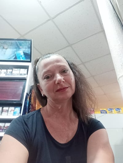 This media file is a selfie taken by a middle-aged woman. She has brown hair pulled back into a ponytail, with some strands loose around her face. She is wearing a dark t-shirt and a silver hoop earring in her left ear. Her makeup includes lipstick.

The setting appears to be an indoor retail environment, possibly a convenience store or a kiosk. Behind her, shelves are visible, stocked with various products, including what appear to be cigarette packs. A television screen is also visible on one of the shelves, displaying an image with blue and white elements. The ceiling is a typical suspended grid ceiling.

The woman's expression is neutral to slightly smiling. There is no indication of the time of day or weather. The location is identified as Bilhorod-Dnistrovskyi, Ukraine. There is some text visible on yellow packaging in the background, but it is difficult to read clearly.