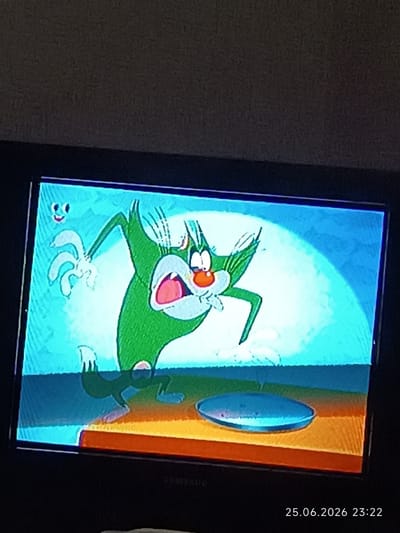 The image displays a cartoon scene on a television screen. A green cat character with a shocked expression, open mouth, and wide eyes is depicted. The cat is shown with its arms raised and a white, abstract object with multiple appendages floating near its left arm. The background is a bright blue with a spotlight effect. In the foreground, on the surface shown on the screen, is a blue, oval-shaped object. The television is positioned in front of a dark, textured surface, likely a wall. A timestamp "25.06.2026 23:22" is overlaid on the bottom right corner of the image, suggesting the photo was taken on this date and time. There is no discernible information within the image or timestamp that would allow for the location in the city of Bolhrad, Ukraine, to be identified.