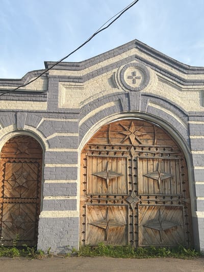 An outdoor, eye-level, vertical shot features a large, ornate wooden gate with two arched entrances. The gate is constructed from weathered wooden planks and decorated with geometric star patterns. The main entrance is a double gate under a large arch, while a smaller, single arched gate is visible to the left. The gate is part of a brick structure with a decorative facade painted in light yellow and gray. Above the gate, the building's roofline is stepped and embellished. A power line runs diagonally across the upper portion of the image, against a clear blue sky. The ground in front of the gate is an asphalt road, with patches of green grass and weeds growing along the base of the structure. The overall impression is one of historical architecture, possibly in a rural or historic district setting. This specific gate is located in the city of Cherdyn, Russia.