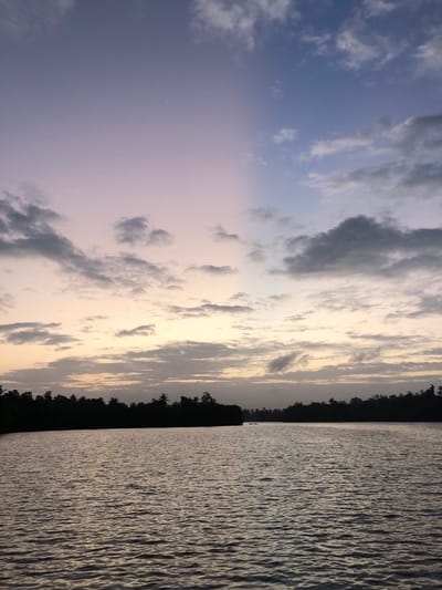 The image depicts a serene water body, likely a lagoon or river, bordering a dense, dark line of trees that forms the silhouette of the Habaraduwa coastline. The sky above is a gradient of soft purples and blues, transitioning into warmer pastel tones near the horizon, indicative of either dawn or dusk. Scattered clouds, varying in shade from light grey to dark, add texture to the sky. The water's surface is rippled, reflecting the muted colors of the sky and creating a shimmering effect. The overall impression is one of tranquility and natural beauty at the edge of Habaraduwa, Sri Lanka.