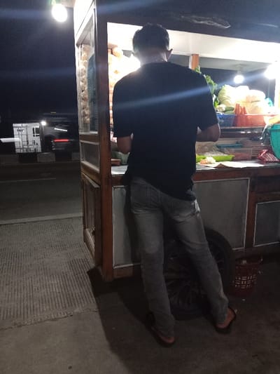 The image shows a man standing at a food stall in Kecamatan Brangsong, Indonesia, located at coordinates -6.947 latitude and 110.228 longitude. The man is wearing a black t-shirt and jeans, and he is looking at something off-camera. He is standing in front of a wooden food stall with a metal frame and a metal roof. There are various food items displayed on the stall, including vegetables, fruit, and other unknown food items. The stall is lit by a single overhead light, and the surrounding area is dark. There is a white van parked in the background, and a black car is parked behind the van. The ground is made of concrete, and there is a sidewalk in front of the stall. The man is seemingly waiting to purchase food from the food stall. There are several unknown food items displayed within the stall. The stall is situated on a busy street.