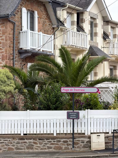 This is a full shot of a street in La Clarté, France, with a white picket fence running along the foreground. Behind the fence, palm trees and various green bushes are visible. A signpost stands in front of the foliage, with two signs attached. The top sign, a white arrow with a pink border, reads "Plage de Trestraou" and points to the right. The bottom sign, a dark gray rectangle with a pink border, reads "Boulevard Aristide BRIAND". In the background, two houses are visible, one with a reddish stone facade and the other with a cream-colored facade. Both houses have balconies with white railings and dark tiled roofs. The sky is overcast.