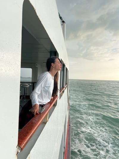 A woman with long dark hair, wearing a white long-sleeved shirt and a red and black plaid skirt, leans out of an open window of a ferry. She is looking out at the sea. Her right hand rests on the wooden railing of the window. A smartphone is placed on the railing near her hand. The railing is wet, suggesting it may have rained recently. The sea is choppy with white foam. The sky is overcast with some light visible on the horizon. The ferry appears to be docked or moving slowly near a landmass, indicated by the distant view of land through the window behind her. The ferry's interior is visible through the window, showing a bar area with stools. The scene takes place on the water, likely en route to or from Don Sak, Thailand, a known ferry hub connecting the mainland to islands like Koh Samui and Koh Phangan.