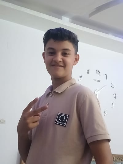 The image shows a young male teenager with dark, curly hair, standing indoors and smiling at the camera. He is wearing a light brown polo shirt with a Moncler logo patch on the left chest. His left hand is raised, with his index and middle fingers extended in a peace sign. Behind him, on a white wall, is a large, modern wall clock with black numerals and hands. The clock appears to be a DIY or sticker clock, with the numbers and hands mounted directly on the wall. The room has white walls and a white ceiling with recessed lighting. The lighting suggests an interior environment. The image does not provide any specific geographical indicators that would place it in the city of Hun, Libya.