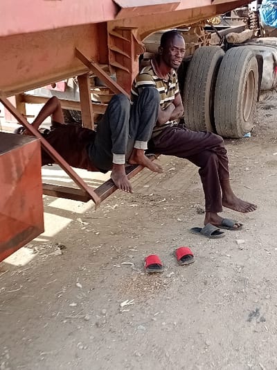 In Sharada, Nigeria, an outdoor scene depicts two adult males resting near a heavy-duty truck trailer. The ground is unpaved, composed of dry dirt and gravel with scattered debris.
One male, wearing a striped polo shirt (brown, black, and light green/beige) and dark trousers, sits barefoot on a horizontal metal support beam of a rusty-orange/red truck trailer. His arms are crossed, and he gazes slightly right with an open-mouth expression; his face shows signs of dust or dirt. Directly below his feet rests a pair of black slide sandals.
Another male is partially visible, lying on his back underneath the same truck trailer. He wears a dark brown/maroon t-shirt and dark trousers. His bare feet are visible, resting on the same metal beam as the sitting man. His face is obscured by the angle and shadows.
In the foreground, to the left of the black sandals, a pair of red slide sandals lies on the ground. In the background, large, dirty truck tires are visible to the right of the sitting man, indicating the presence of a full truck or another trailer section. Natural daylight illuminates the scene, casting shadows from the truck and individuals.