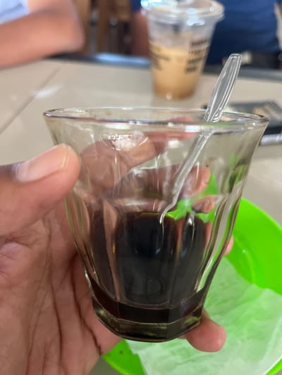 In Panteraya, Indonesia, a close-up shot captures a hand holding a small, clear glass tumbler filled with dark liquid, likely coffee, and a clear plastic spoon. The tumbler's faceted sides reflect light and the surrounding environment, including a vibrant green plate underneath, which is lined with white paper. In the background, out of focus, a person's arms are visible, suggesting a casual setting such as a cafe or restaurant. Another out-of-focus disposable cup with a straw, containing a light-colored beverage, is also present in the background. The lighting suggests a well-lit indoor or shaded outdoor environment.