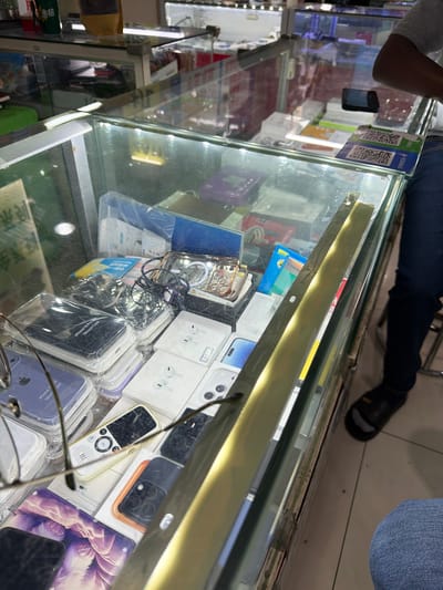Captured from an elevated, angled perspective, the photograph presents a view into a glass display case, likely situated in a commercial area of Xuanwu District, China. Within the case, a diverse assortment of mobile phone accessories is arranged. These include cases in various colors, possibly for iPhone 16 and iPhone 17 models, as indicated by labels in English and Chinese. Additionally, there are boxes that appear to contain wireless earbuds or earphones, potentially manufactured by Apple given the packaging.
In the periphery of the frame, elements of the surrounding environment are visible. The legs of individuals, clad in denim jeans, suggest customers or shopkeepers are present. The foot of a man in black slippers is seen as well. These elements imply a bustling commercial atmosphere, possibly within a small shop or market stall where mobile phone accessories are sold.
