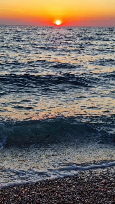 This is a vertical, wide-angle shot of a coastline at sunset. The sun, a bright orange orb, is setting on the left side of the frame, casting a warm, golden glow across the sky. The water, in the center of the image, is a deep blue with white-capped waves reflecting the sunset's colors. The waves appear to be gently rolling towards the shore, creating a frothy white line where they meet the beach. The beach itself is made up of small, dark pebbles, some of which are illuminated by the setting sun. The overall mood of the image is peaceful and serene, with the warm colors of the sunset creating a sense of tranquility. The location appears to be a beach, likely in Zadar, Croatia, given the context. The time of day is sunset.