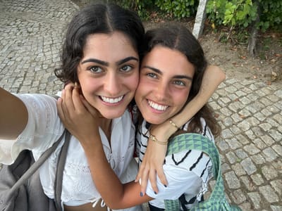 Two young women are taking a selfie outdoors in Felgueiras, Portugal. The woman on the left, with dark curly hair and brown eyes, is wearing a white top and has her arm around the other woman. The woman on the right, with dark brown hair and green eyes, is wearing a white top with black and green geometric patterns and has her arm around the first woman. They are both smiling and have their faces close together. The background shows a cobblestone path and greenery, suggesting a park or garden setting.
