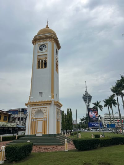 The image shows the Alor Setar Tower and the Clock Tower in Alor Setar, Malaysia, on an overcast day. In the foreground, the white and gold clock tower stands prominently. It features a golden dome, clock faces on each side, and decorative arches at its base. Steps lead up to the tower, which is surrounded by a manicured lawn and low hedges. A red brick pathway surrounds the base of the clock tower. To the right of the clock tower, palm trees line the street. In the background, the Alor Setar Tower, a tall, slender structure with a unique design, is visible. A large billboard displays advertisements and text, including "SELAMAT DATANG KE ALOR SETAR." Cars are seen driving on the road in front of the tower and billboard. Buildings, including one with a yellow facade, are present to the left of the clock tower. The sky is filled with heavy gray clouds. There are no people visible in the image.