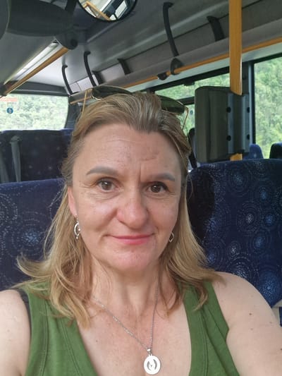 This is a selfie taken from a seated position inside a bus. The subject is a woman with blonde hair and light skin, wearing a green tank top and a silver necklace with a pendant. She is looking directly at the camera with a slight smile. Sunglasses are perched on her head. The background shows the interior of the bus, including blue seats with a patterned design, and windows through which trees and foliage are visible. The overhead interior of the bus is also visible, with yellow poles and handles. The location is not specified as Verrès, Italy.