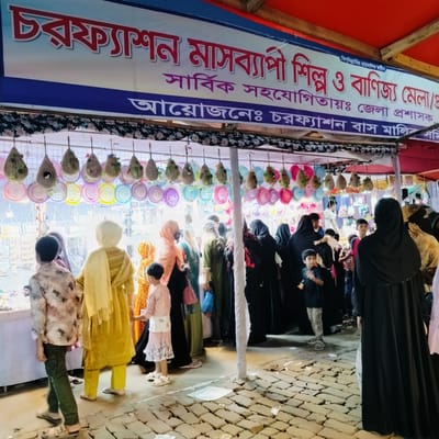 The image depicts a bustling outdoor market or fair stall at night, likely in Charfasson, Bangladesh, given the prominent Bengali text on the banner. Numerous people, including women in hijabs and abayas, men, and children, are seen browsing merchandise. The scene is illuminated by artificial lighting, highlighting the vibrant colors of the items on display.

The stall features various goods, with decorative items hanging above, resembling small bird nests made of natural fibers and colorful woven hats or plates. Below these, shelves or tables are filled with smaller items, possibly jewelry or accessories, which shoppers are examining. The ground is paved with bricks or pavers, showing some wear and debris typical of a busy marketplace.

A large banner spans the top of the stall with Bengali text. The visible text, as transcribed by OCR, reads:
"বিসমিল্লাহির" (In the name of Allah, the Most Gracious, the Most Merciful)
"চরফ্যাশন মাসব্যাপী শিল্প ও বাণিজ্য মেলা/" (Charfasson Month-long Industry and Commerce Fair/)
"সার্বিক সহযোগিতায়ঃ জেলা প্রশাসক" (With overall cooperation: Deputy Commissioner)
"আয়োজনেঃ চরফ্যাশন বাস মালিক সমি" (Organized by: Charfasson Bus Owners Association)

This indicates the event is a month-long industrial and commercial fair in Charfasson, organized by the Charfasson Bus Owners Association with the cooperation of the Deputy Commissioner. The general atmosphere suggests active commerce and community engagement at the fair.