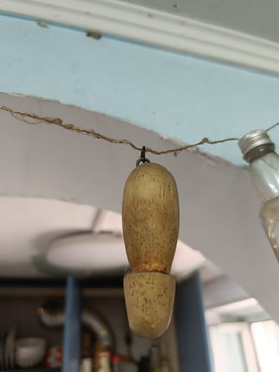 A close-up, low-angle shot shows a wooden object hanging from a thin, gold-colored wire. The wooden object is roughly cylindrical with a rounded top and a narrower, flared bottom. It appears to be a decorative element or a part of a larger item. The wire is strung across an archway. To the right of the wooden object, a glass bottle with a silver cap is also hanging from the wire, appearing to be part of the same decorative string of objects. The background is out of focus, showing a light blue wall and a white ceiling. Below the archway, a shelf unit with various items, possibly in a kitchen or storage area, is visible. There is no indication of the location being in Taraz city administration, Kazakhstan.