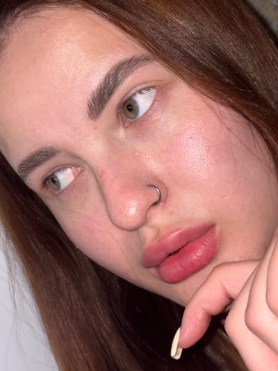 Close-up of a woman's face rotated at an angle. She has light skin, brown hair, and green eyes. A silver ring is visible in her left nostril. Her right hand is near her lips.
Port-Katon, Russia is not discernible in the image.