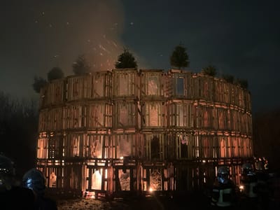 A large, cylindrical structure of wooden pallets burns fiercely at night. Flames are visible through the open spaces of the pallets. Sparks fly upwards. Several small trees or shrubs are placed atop the structure. Dark silhouettes of firefighters in protective gear stand in the foreground. The background is dark, with trees visible to the side and a dark sky above.