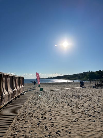 The image depicts a sunny day at a sandy beach with a clear blue sky. The sun is bright and positioned in the upper left quadrant of the frame, casting long shadows across the sand. A wooden boardwalk runs along the beach, and several people are visible walking on it and on the sand. A red flag with white lettering is planted in the sand near the water's edge. The text on the flag reads "COFFEE LIKE." In the background, there is a line of trees bordering the beach, and beyond that, a body of water that appears calm and reflects the sunlight. The overall scene evokes a relaxed and pleasant atmosphere, suggesting a typical day at the beach during warm weather. The time of day appears to be late afternoon, given the length of the shadows. The location is Volzhskiy Utes, Russia.
