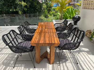 The image shows an outdoor dining area in Buduk, Indonesia. A rectangular wooden table with a unique, geometric base is surrounded by eight black wicker chairs with gray cushions. The scene is bathed in sunlight, casting striped shadows across the white tiled floor. Behind the dining set, a glass barrier separates the area from lush greenery, including various plants and trees. A black barbecue grill stands to the right of the table near a white wall, adding to the outdoor setting.