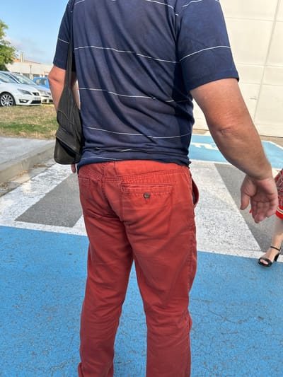 An individual wearing a navy blue horizontally striped t-shirt and red trousers stands on a street in Cerdanyola del Vallès, Spain. The ground is a textured surface with blue and white painted markings, indicating a crosswalk. In the background, several parked cars are visible, including a white sedan and a blue vehicle. The individual is partially obscured from view from the waist up, with a black bag slung over their left shoulder. A portion of another person's legs and feet, wearing black high-heeled sandals, are visible to the right of the frame. The lighting suggests it is daytime, with shadows cast on the ground.