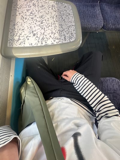 The image shows a person sitting on a train seat, likely in the Folkestone and Hythe District, United Kingdom, given the typical train interiors found in the region. The person is wearing a black and white striped long-sleeved shirt and black trousers. They are holding a pair of sunglasses in their right hand. In front of the person is a foldable tray table with a speckled pattern. A khaki-colored cushion is resting on the person's lap. The train seats are upholstered in a dark blue fabric with a textured pattern. The floor of the train appears to be carpeted in a dark color. The overall environment suggests a journey on public transportation.